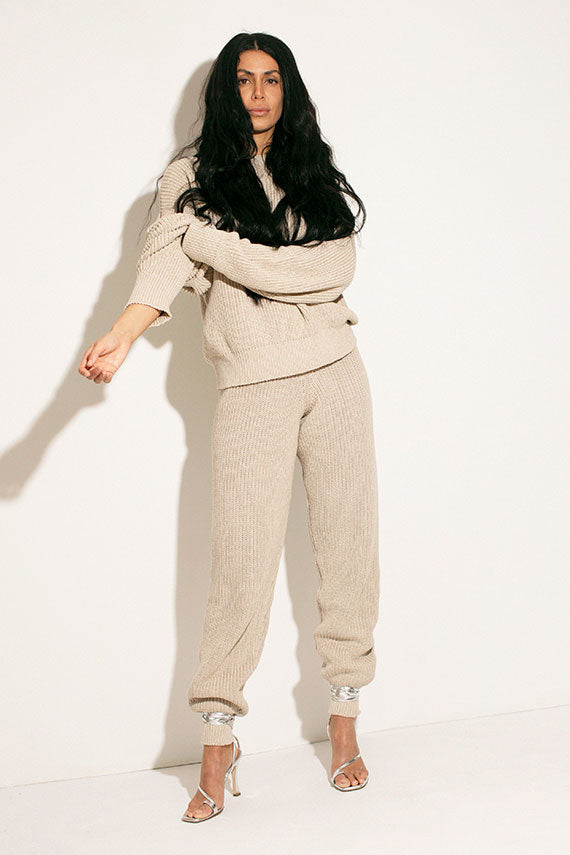 Model pulls up the sleeves of the Baserange sand melange pullover sweater, wearing matching pants and Brother Vellies silver heels